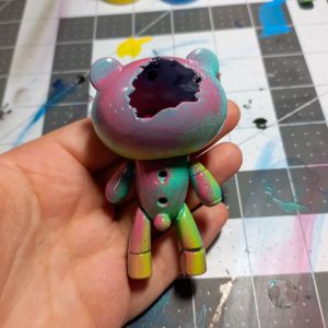 Shows a petit beargguy that is colorful and sickly. It is smooth in the front. The colors are aqua blue, pink, and green. The skull is crushed in on the back.